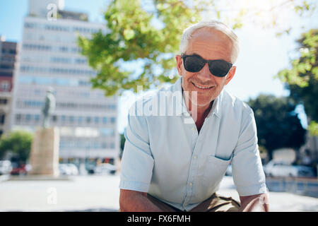 Ritratto di felice senior uomo seduto al di fuori della città. Uomo maturo con occhiali da sole all'aperto su un giorno d'estate. Foto Stock