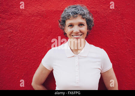 Ritratto di donna senior in piedi contro lo sfondo di colore rosso. Sorridente metà femmina adulta contro la parete rossa. Foto Stock