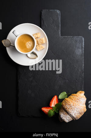 Tazza di caffè e un croissant con fresco strawbwrries su nero ardesia scheda di pietra su sfondo scuro. Vista superiore, spazio di copia Foto Stock