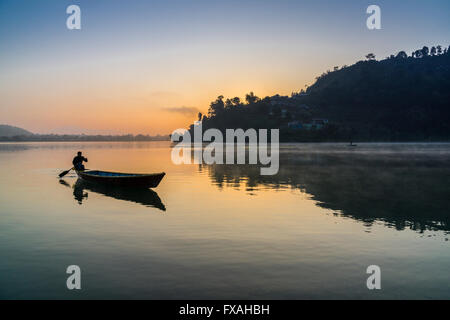 L'uomo paddling in barca su lago Phewa presso sunrise, Pokhara in distanza, nebbia, Anadu Palpari, Kaski, Nepal Foto Stock