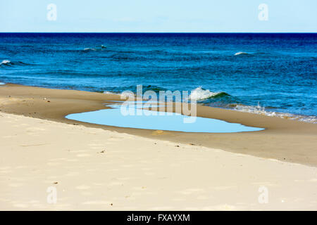 Pozza d'acqua di mare poco profondo sulla spiaggia sabbiosa con il mare aperto in background. Piccole onde alimentare la pozzanghera con nuova acqua. Foto Stock