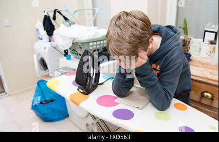 White ragazzo adolescente con la testa tra le mani appoggiate su di un macchie asse da stiro nel ripostiglio con lavanderia e lavaggio intorno a lui Foto Stock