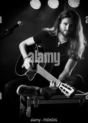 Foto di un giovane uomo con i capelli lunghi e la barba suonando una chitarra acustica sul palco con luci e atmosfera da concerto. Foto Stock