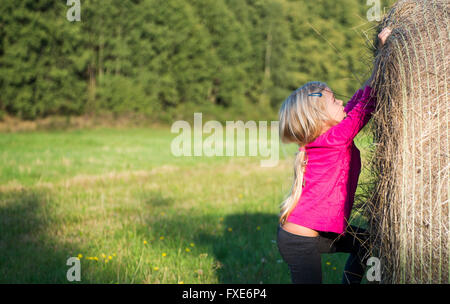 Bambino ragazza bionda da paglia balla di fieno in campo, prato, giocare i bambini attività estive Foto Stock