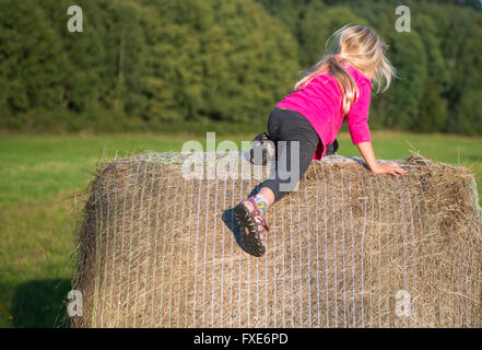 Bambino ragazza bionda da paglia balla di fieno in campo, prato, giocare i bambini attività estive Foto Stock