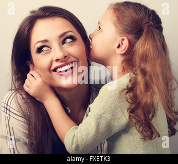 Felice carina ragazza di capretto whispering il segreto per la sua madre sorridente in orecchio con faccia di divertimento. Tonica closeup ritratto Foto Stock