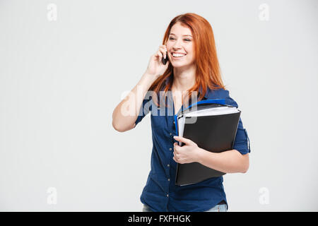 Allegro grazioso redhead giovane donna con cartelle parlando al cellulare su sfondo bianco Foto Stock