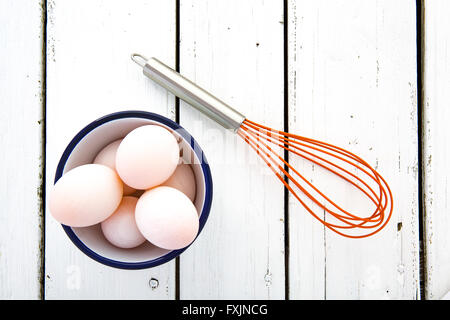 Le uova in una ciotola di ceramica con una frusta su un bianco rustico in legno dipinto table top shot dal di sopra Foto Stock