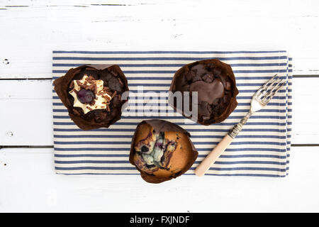 Tre deliziosi muffin al cioccolato su un blu e bianco spogliato igienico con una forcella su un bianco tavolo rustico Foto Stock