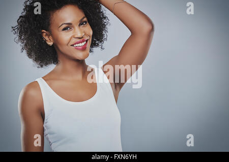 Unico sorridente bella donna afro-americana che indossa senza maniche canottiera bianca con mano verso la testa sulla copia grigio spac Foto Stock