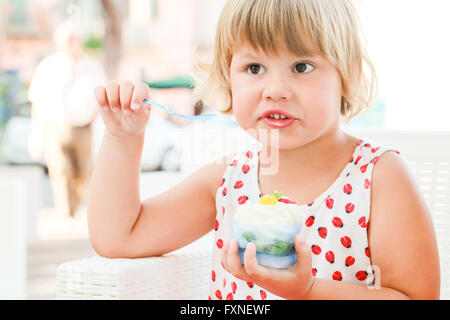 Carino biondo Caucasian Baby girl mangia frozen yogurt con gelato e frutta, closeup ritratto all'aperto con naturale alta paglierino chiave Foto Stock