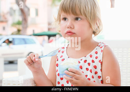 Carino biondo Caucasian Baby girl mangia lo yogurt con gelato e frutta, chiudere fino all'aperto verticale con naturale alta luce chiave Foto Stock