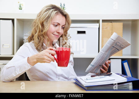 Sorridente business donna nel suo ufficio leggendo il giornale mentre si beve il caffè Foto Stock