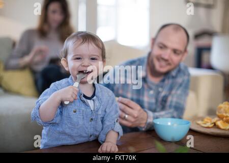 Baby boy alimentare se stesso con cucchiaio guardando sorridente della fotocamera Foto Stock