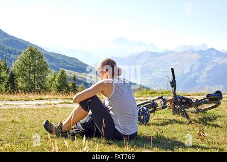 Femmina mountain biker seduta nel paesaggio di montagna la lettura di testi di smartphone, Valle d'Aosta, Aosta, Italia Foto Stock