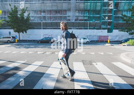 Giovane uomo attraversamento attraversamento pedonale con lo skateboard Foto Stock