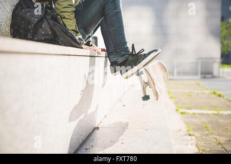 Cintura vista verso il basso del giovane maschio urban skate boarder seduto sul muro di capovolgimento di skateboard con piedini Foto Stock