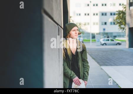 Giovane maschio guidatore di skateboard urbano appoggiata contro la parete ascoltando musica auricolare Foto Stock