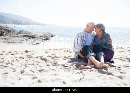 Coppia senior seduto sulla spiaggia, avvolgente, faccia a faccia sorridente Foto Stock