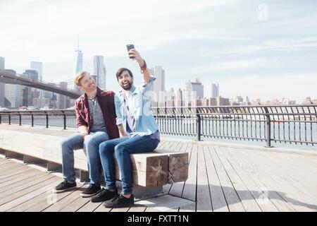 Voce maschile giovane seduto sul fiume dal Ponte di Brooklyn prendendo selfie smartphone, New York, Stati Uniti d'America Foto Stock