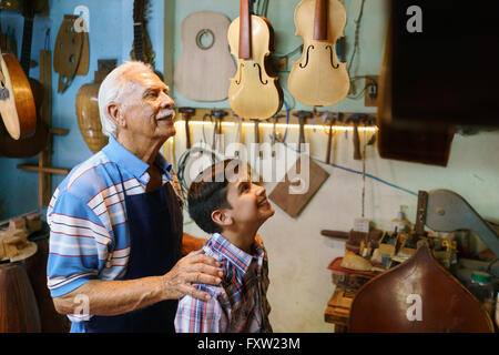 Piccola azienda di famiglia e tradizioni: il nonno vecchio con il nipote in liutaio shop. L'artigiano senior avvolge il ragazzo e mostra hi Foto Stock