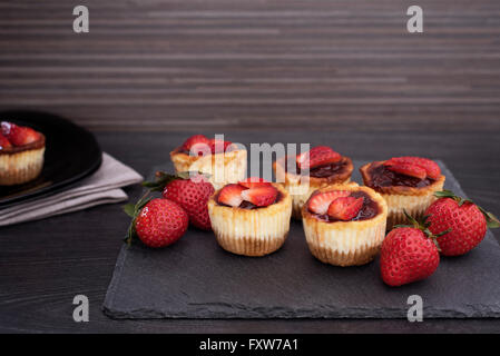 Mini Strawberry Cheesecake in forme di muffin. Servito in pietra nera altopiano con fragole fresche. In legno nero e sfondo. Foto Stock