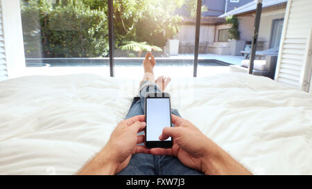 Primo piano immagine della mano umana tenendo il telefono cellulare con schermo vuoto sul letto. POV shot di uomo disteso sul letto tramite smart phone. Foto Stock