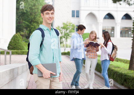 International Student in college con zaino in possesso di un libro e sorridente in avanti con lo sfondo dei suoi amici insieme al campus Foto Stock