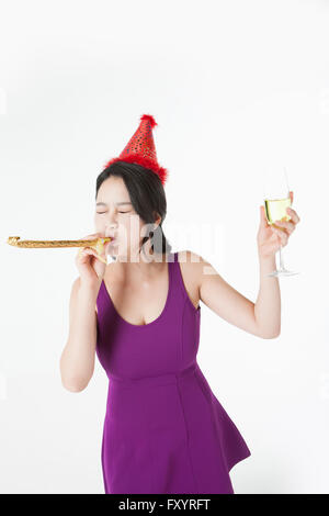 Giovane donna soffia un corno di partito con in mano un bicchiere di champagne chiudendo gli occhi Foto Stock