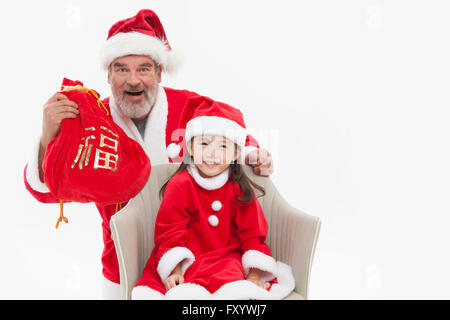 Sorridente Santa Claus che mostra un sacco di fortuna e un sorridente ragazza seduta su una sedia con lo sguardo sul lato anteriore Foto Stock