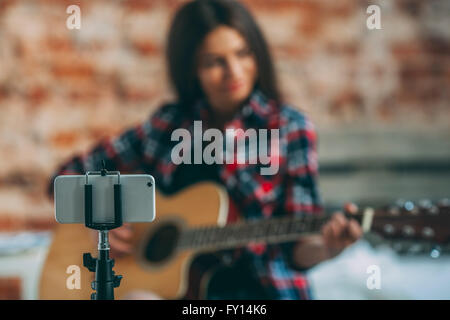 Smart phone in monopiede con la donna a suonare la chitarra in background a casa Foto Stock