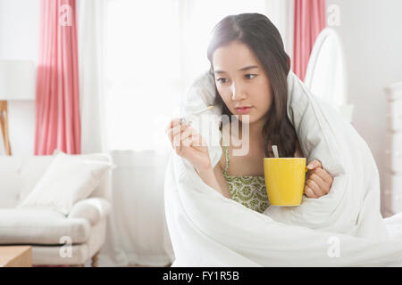 Ritratto di giovane donna malata in coperta holding e guardando il termometro con una tazza Foto Stock