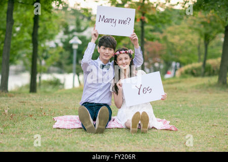 Sposa e lo sposo tenendo un messaggio di marry me e sì sia seduto su un'erba Foto Stock