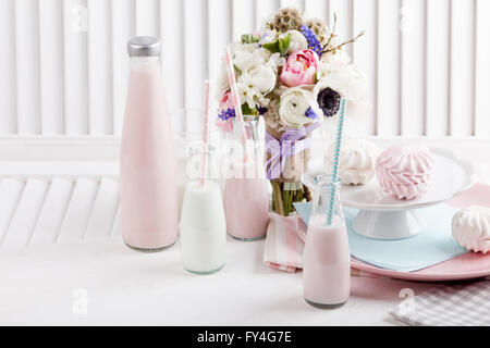 Quattro bottiglie di frappè, tre con cannucce e bicchiere di latte e le piastre con rosa e bianco meringhe e panno sul legno bianco Foto Stock