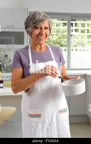 Donna anziana la cottura in cucina in piedi battendo gli ingredienti in un recipiente di miscelazione mentre guardando la telecamera con un sorriso Foto Stock