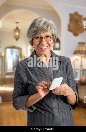 Nonna con gli occhiali in piedi al chiuso nella sua casa utilizzando un telefono intelligente Foto Stock