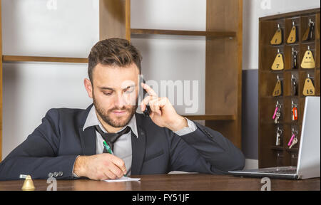 Ritratto di un receptionist prendere nota su un pezzo di carta mentre si è al telefono alla sua scrivania in un piccolo ostello. Foto Stock