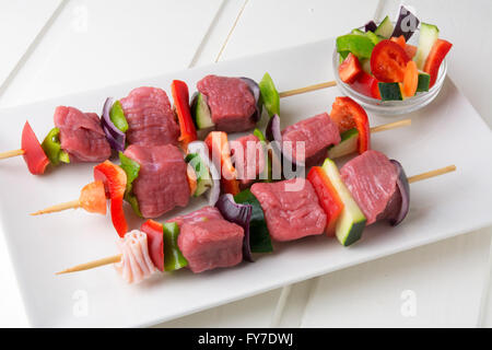 Raw spiedini di carne con verdure fresche sulla piastra rettangolare bianco e tavolo in legno Foto Stock