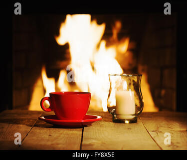 Red tazza di caffè o di tè e la candela sul tavolo di legno vicino al camino. Inverno e vacanze di Natale concetto. Foto Stock