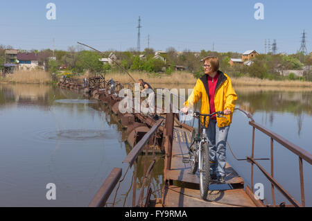 Dnepropetrovsk oblast, Ucraina - 23 Aprile 2016:donna matura camminando con la bicicletta su una chiatta-ponte sul fiume piccolo Sura Foto Stock