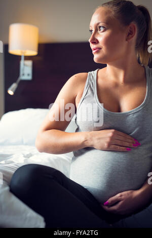 Donna incinta si siede sul letto con la sua mano a pancia Foto Stock