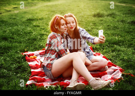 Due giovani ragazze Tanga divertendosi e scattare foto (rendendo Selfie) sullo smartphone al picnic in estate il parco al tramonto. Foto Stock