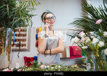 Allegro carino donna giovane fioraio in piedi e parlando al telefono cellulare in negozio di fiori Foto Stock