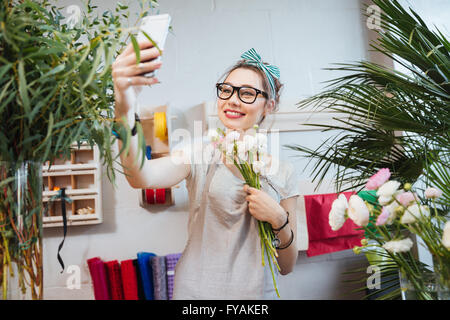 Felice piuttosto giovane donna fioraio tenendo selfie con lo smartphone nel negozio di fiori Foto Stock