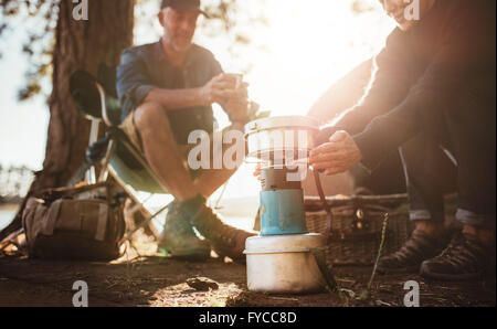 Giovane seduto presso il campeggio in una giornata di sole, con la donna riscaldando le mani sulla stufa di camp. Foto Stock