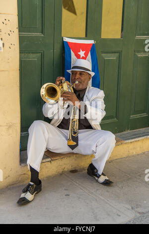 Un musicista cubano svolge la sua tromba, indossando una tromba Tie collo. Seduta su un gradino con una bandiera cubana dietro. Vecchia Havana, Cuba. Foto Stock