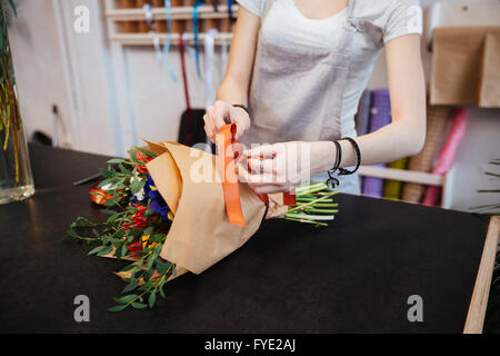 Primo piano delle mani del giovane donna fioraio rendendo bouquet di fiori con nastro rosso in negozio Foto Stock