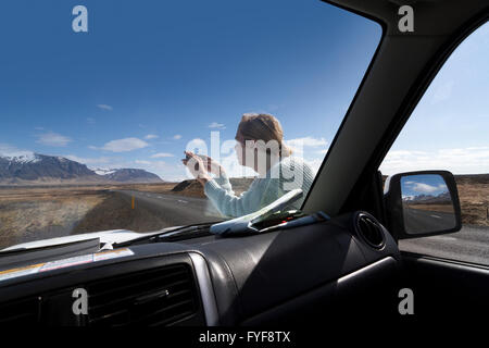 Immagine orizzontale di una donna bionda fare foto con il suo telefono mentre prendendo una pausa sulla strada di circonvallazione in Islanda, fotografato Foto Stock