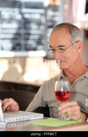 Grigio uomo dai capelli utilizzando portatile mentre bere un bicchiere di vino Foto Stock