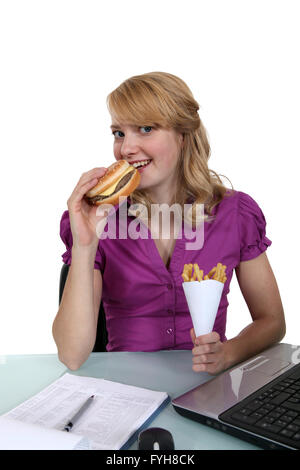 Donna di mangiare un cheeseburger e patatine fritte alla sua scrivania Foto Stock